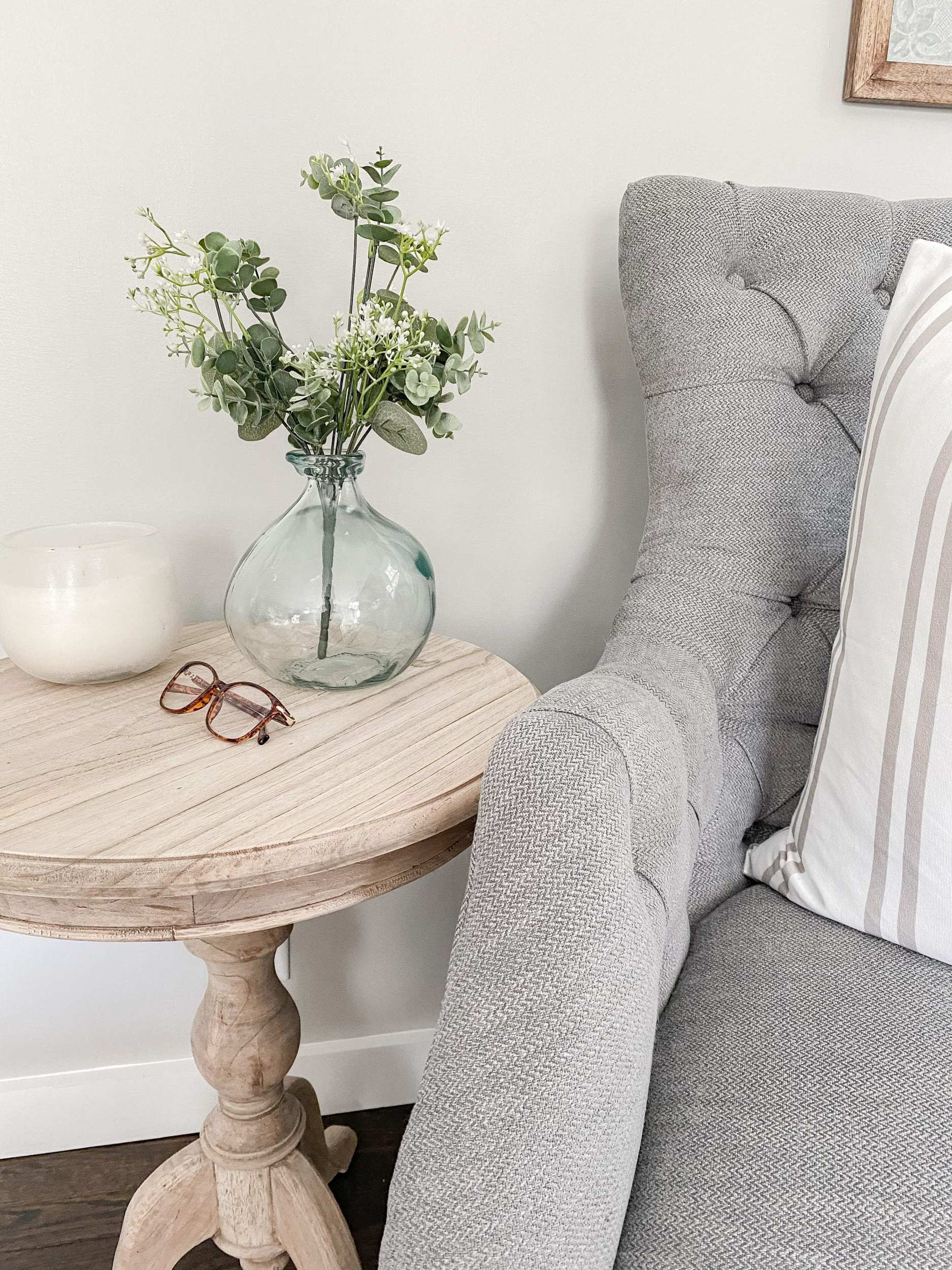 grey tufted armchair with unfinished wood side table, candle, demijohn, and reading glasses