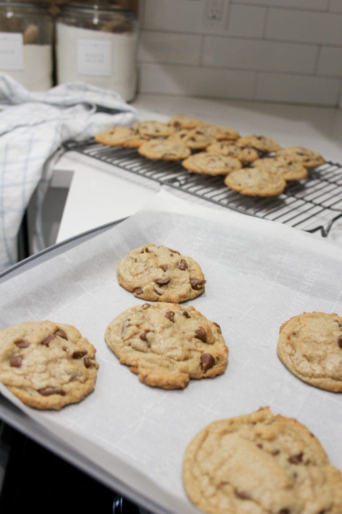 Finished chocolate chip cookies cooling on a rack with fresh cookies just out of the oven on a parchment lined silver baking sheet. 