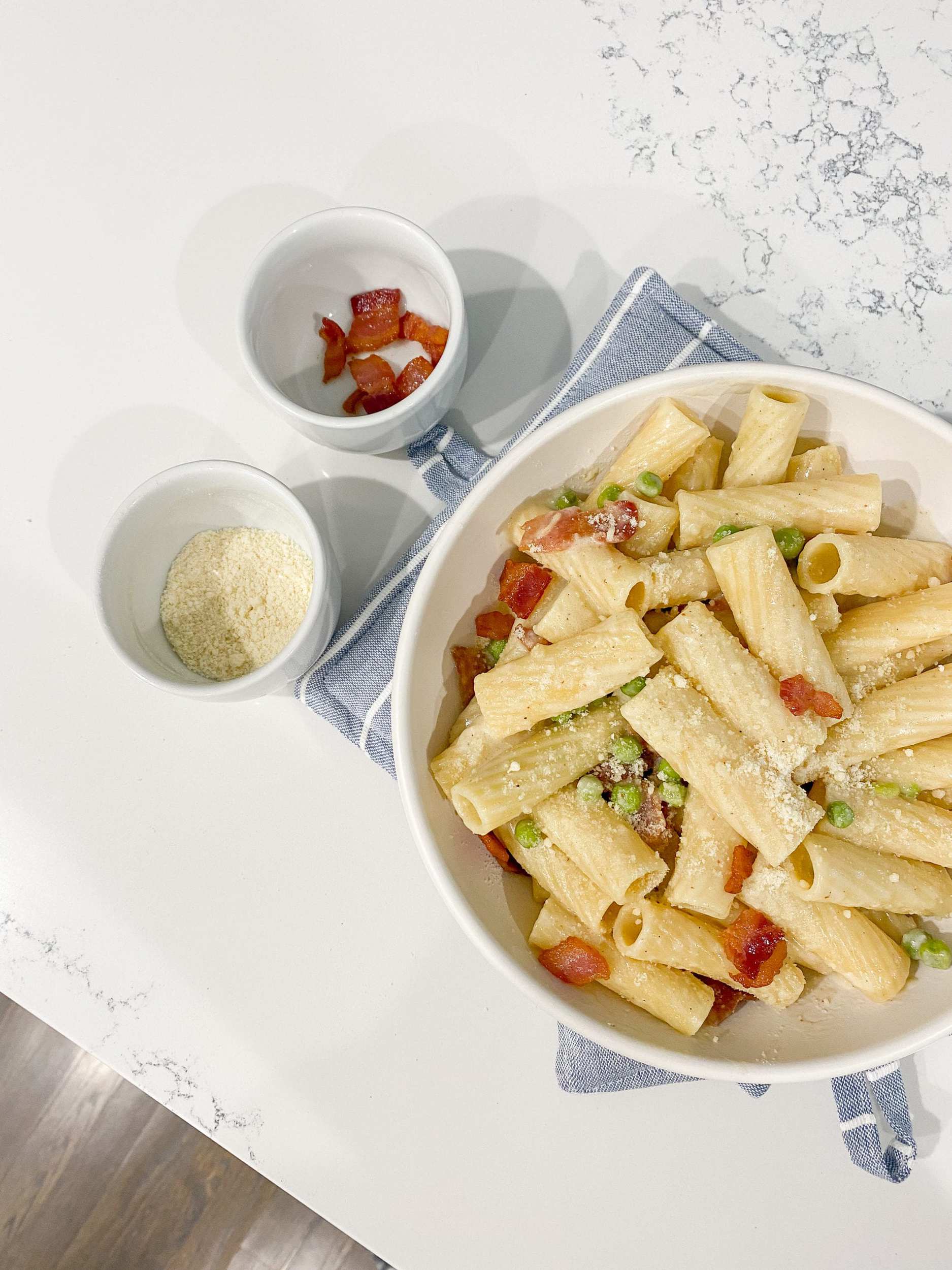 Rigatoni pasta in a matte white pasta bowl with parmesan cheese, bacon, and peas. 