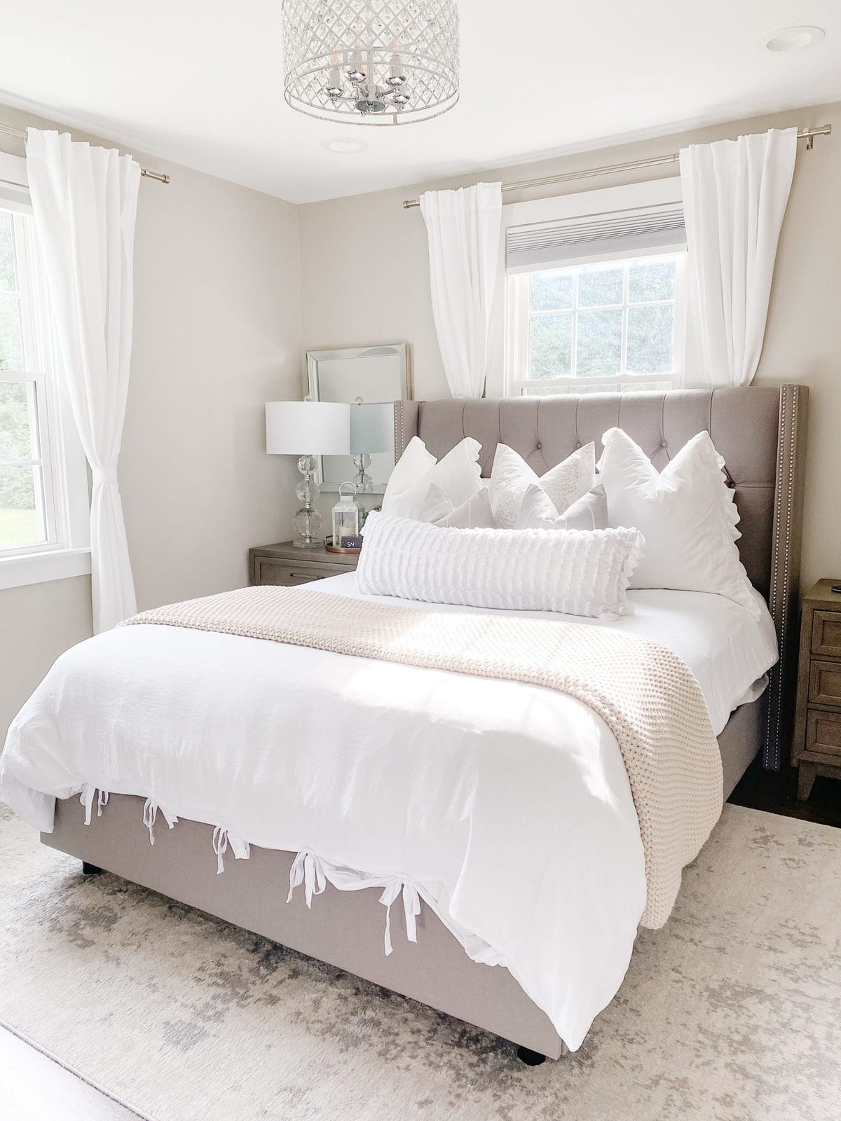 Farmhouse glam bedroom with textured cream knit blanket, white textured lumbar pillow, grey and white striped pillows, white ruffled euro pillows, grey tufted headboard, grey nightstands, crystal nightstand lamps, mirrors