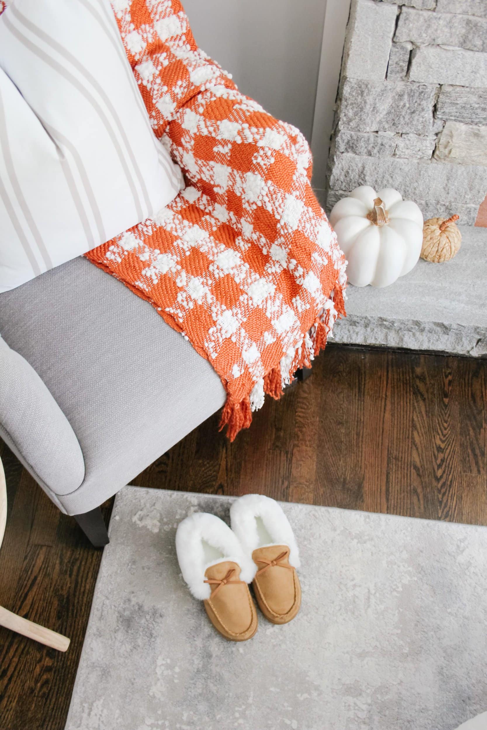 Tufted grey reading chair with orange and white buffalo check blanket, white pumpkins, and tan moccasins. Cozy fall blanket. 