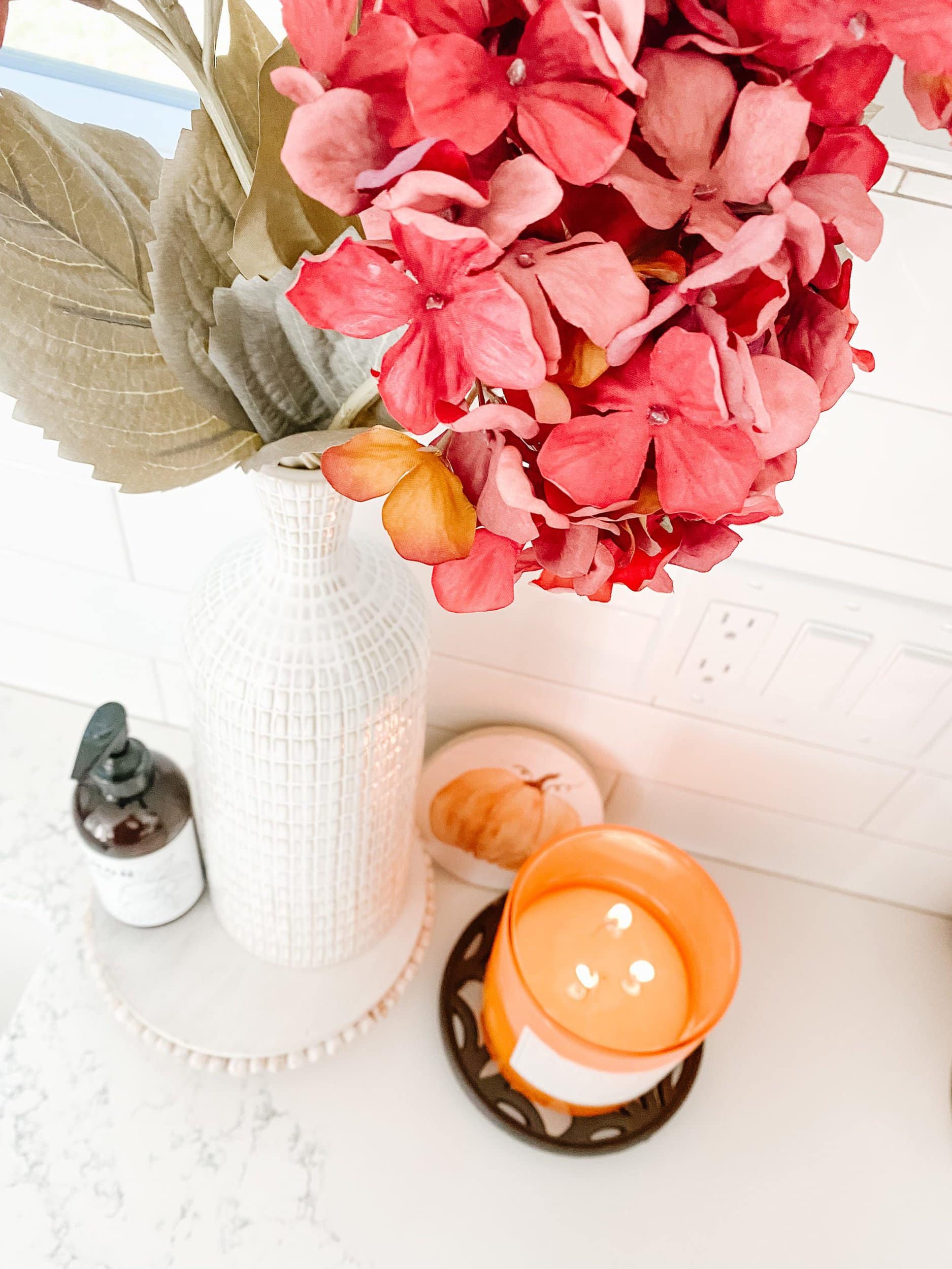 Faux berry colored hydrangea stems, magnolia home soap, ceramic vase, white riser, and pumpkin candle burning on a white quartz countertop