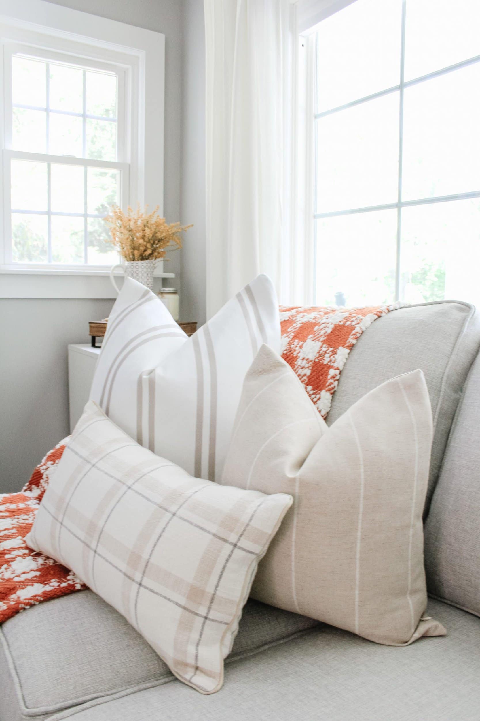 Orange and white plaid buffalo check blanket with white and taupe striped pillow, tan and white striped pillow, and grey, tan, and white plaid pillow on grey couch 