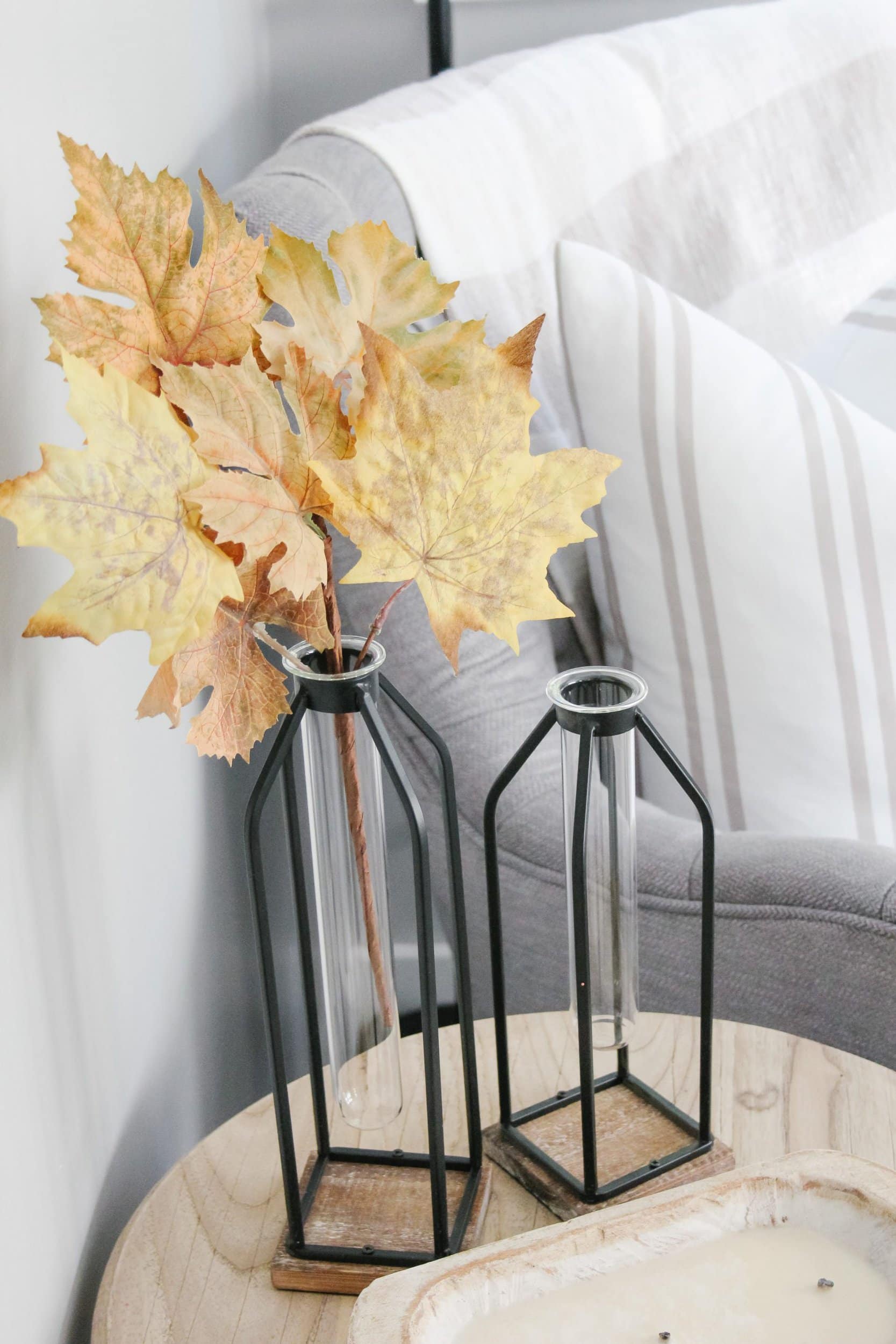 Clear and black test tube bottle vases with faux maple leaves and a tufted grey reading chair and white and taupe striped pillow
