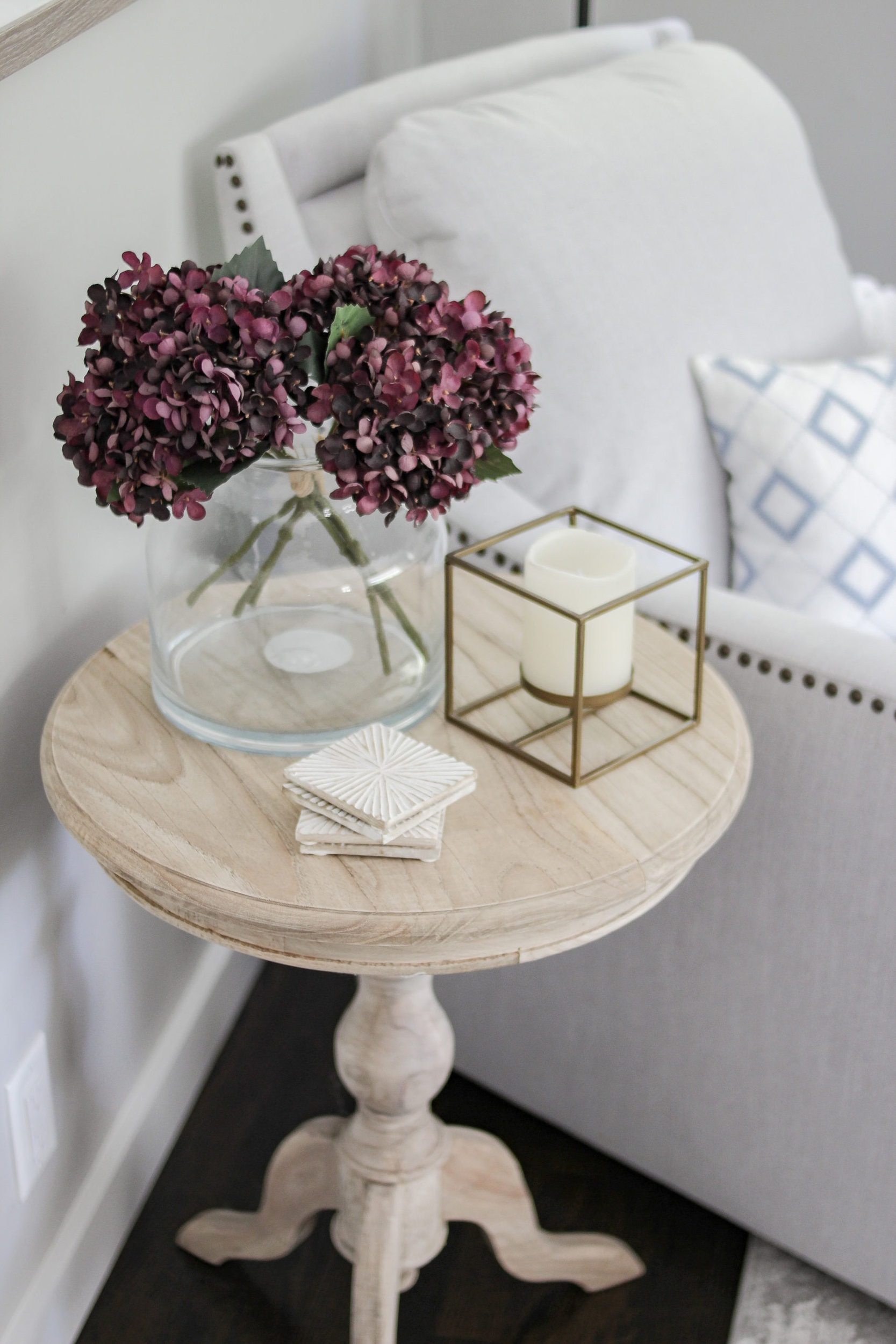 Third & Main starburst mango wood acacia coasters on an unfinished round side table with a studio mcgee vase filled with purple hydrangeas, a geometrical candle holder, and a grey grommet detailed swivel chair