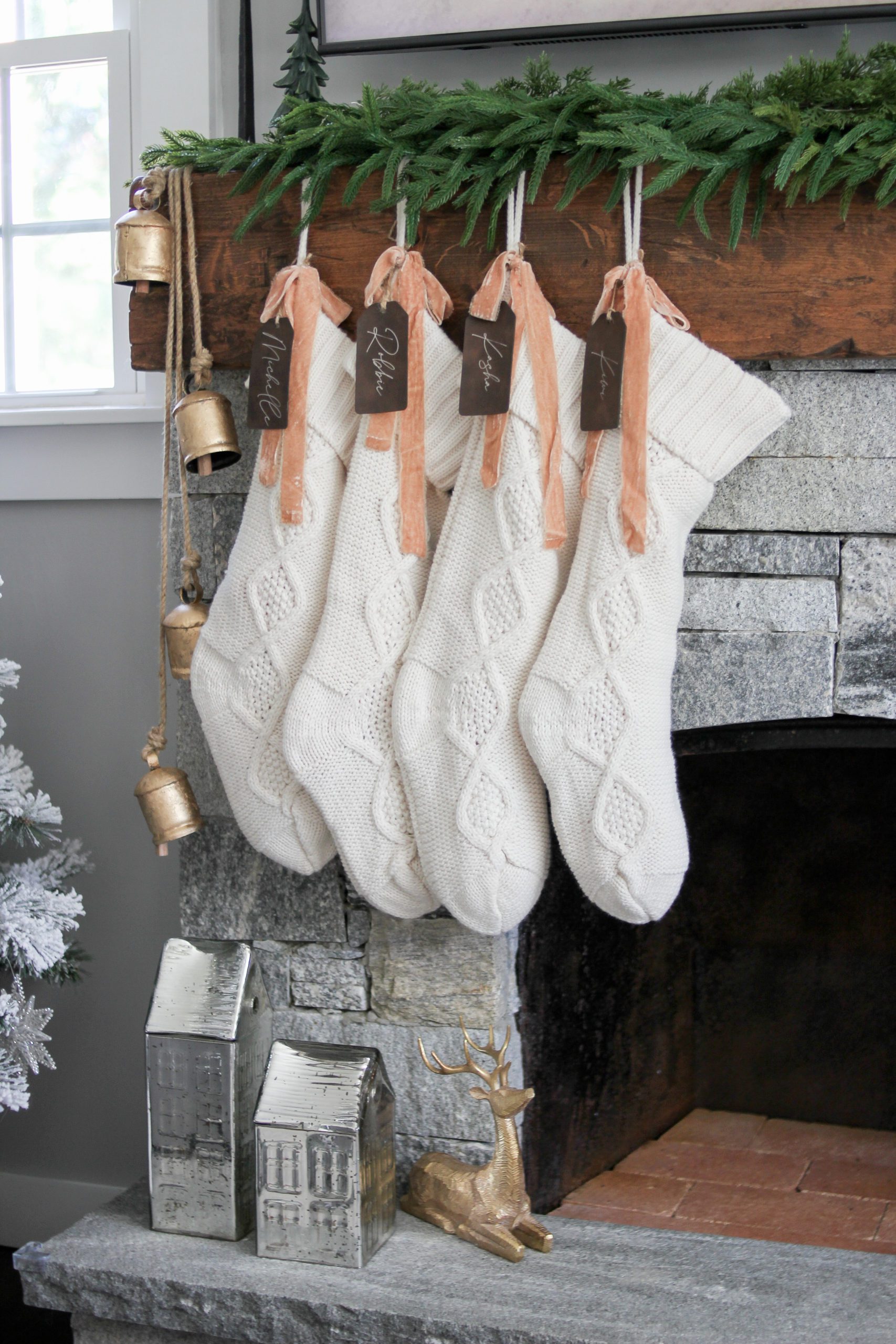 white ivory knit stockings hanging on a grey stone fireplace with fawn ribbon, leather stocking tags, brass hanging bells, and cedar real touch garland