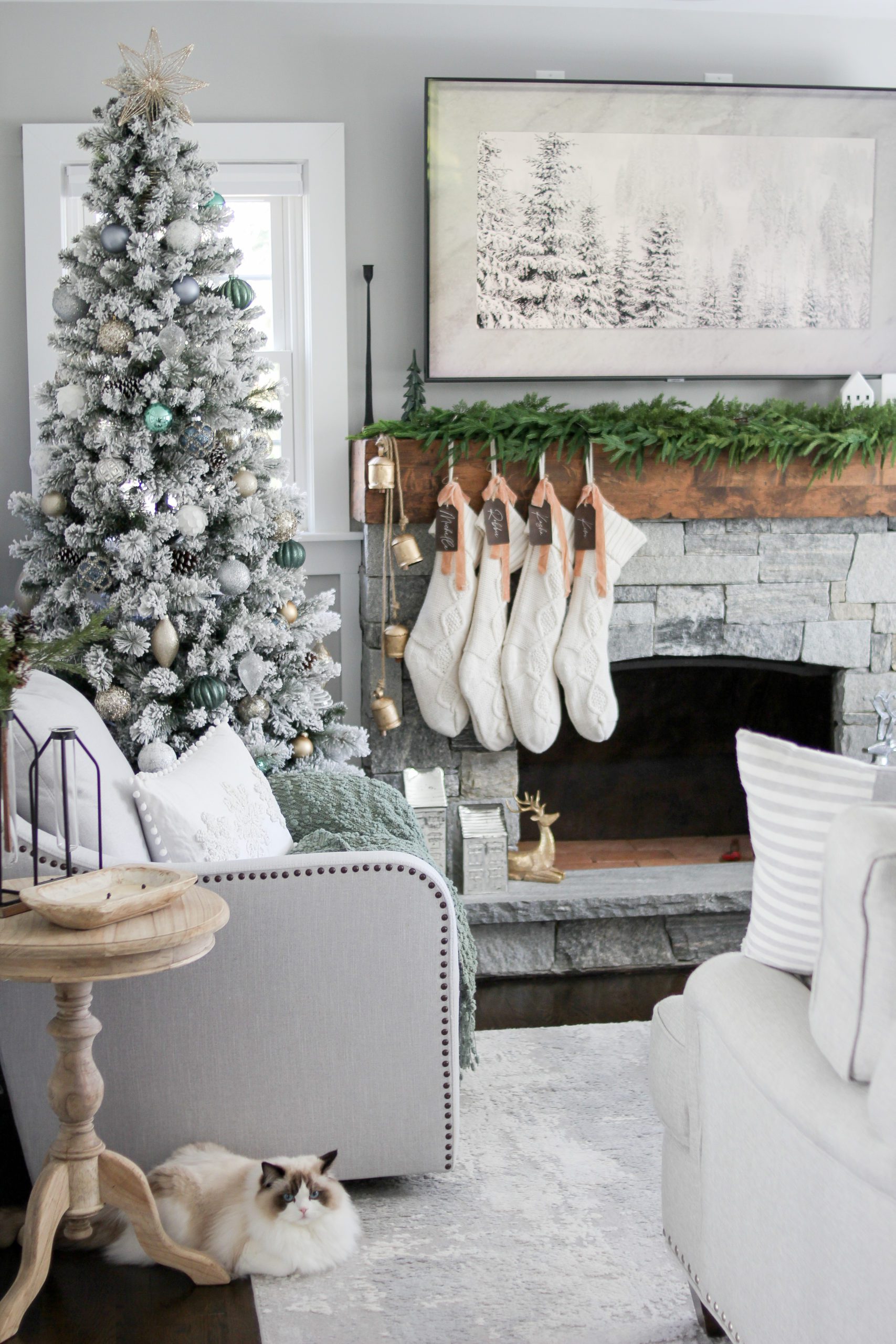 Flocked narrow christmas tree for small spaces decorated with green, blue, champage, gold, and silver ornaments with a grey stone fireplace, wooden mantle, cedar garland, white knit stockings, leather stocking tags, fawn ribbon, and brass hanging bells 