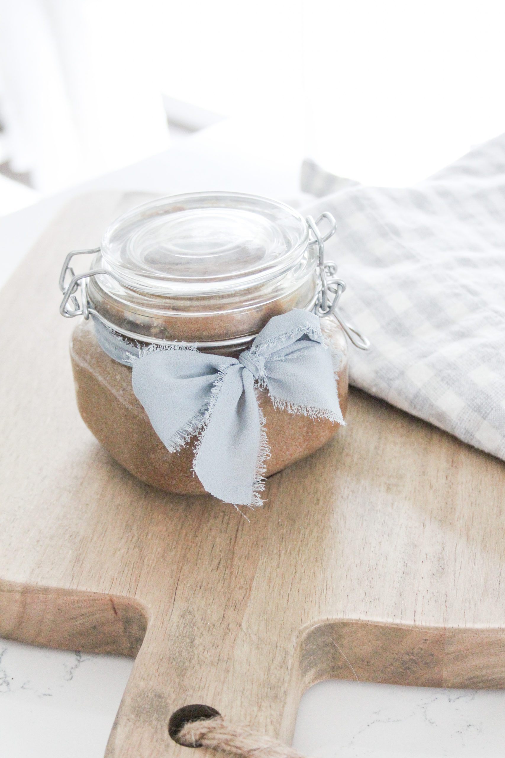 vanilla and honey brown sugar scrub in a mason jar clamp shit container on a cutting board with a white and blue checkered apron in a farmhouse kitchen